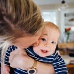 Mum kisses smiling baby in navy striped shirt, surrounded by newborn essentials and baby feeding products indoors.
