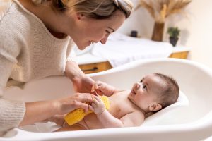 Mum bathes smiling baby in white tub with yellow sponge, surrounded by newborn essentials and soft decor.