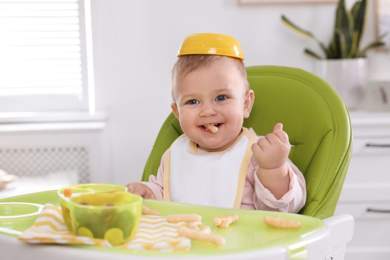 Smiling baby in a green high chair, yellow bowl on head, enjoying Aussie baby feeding products with snacks scattered on tray.