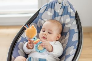 Curious baby in a blue and grey seat holding an orange and blue rattle, exploring newborn essentials.