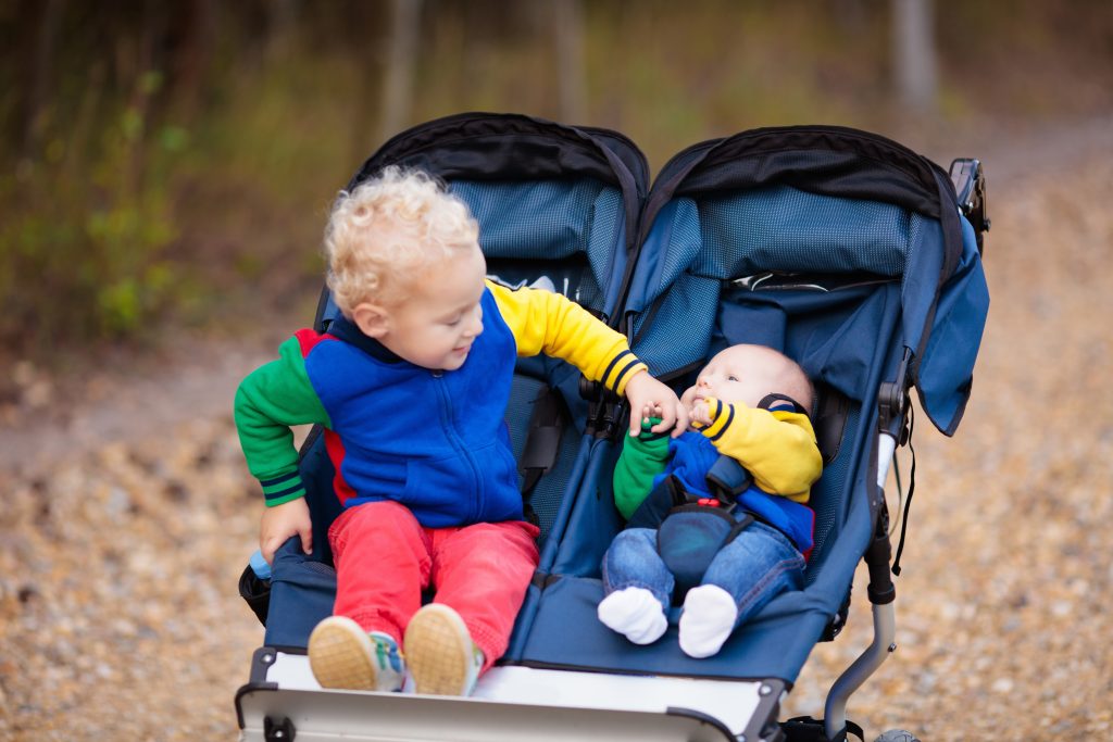 Toddler and baby in a double stroller outdoors, toddler touches baby's hand; enjoying fresh air in colourful jackets.