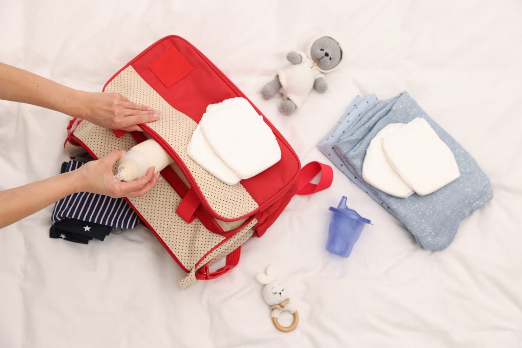 Packing a red diaper bag with newborn essentials on a white baby play mat, featuring sippy cup, bottle and plush toy.