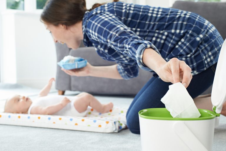 Mum changes baby on patterned play mat, tossing a used wipe in the bin and holding clean wipes; showing newborn essentials.