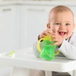 Smiling baby in high chair holding green sippy cup, with pacifier on tray in a white kitchen—ideal for baby product guide.