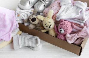 Box with folded baby clothes, socks, toys, pink bow and silver baby shoes—newborn essentials for Aussie mums.