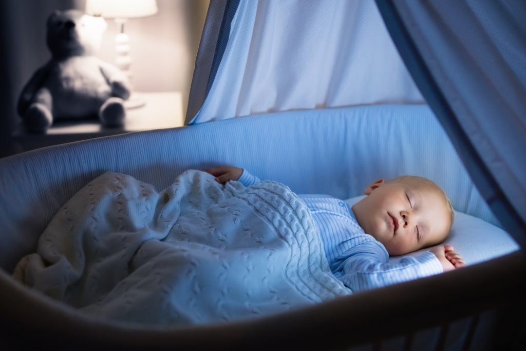 Sleeping baby in crib under soft blue blanket, with newborn essentials like a teddy bear and lamp in the background.