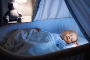 Sleeping baby in crib under soft blue blanket, with newborn essentials like a teddy bear and lamp in the background.
