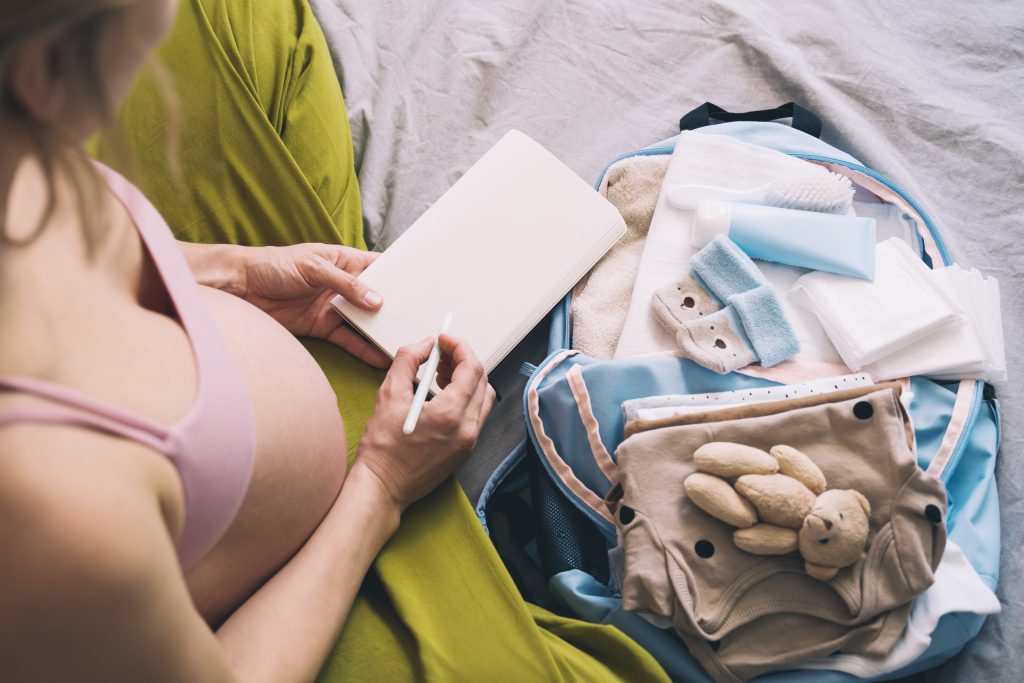 Pregnant woman in pink bra and green pants writes beside a hospital bag packed with newborn essentials, baby clothes, and toys.