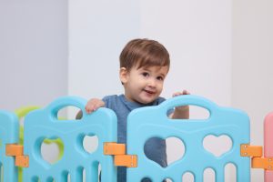 Smiling toddler in blue shirt stands behind a light blue play fence with heart cutouts. Featured in our baby products Australia guide.