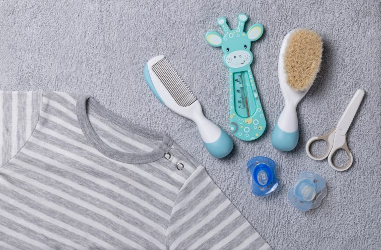 Newborn essentials: striped onesie, baby brush, comb, thermometer, scissors and pacifiers on a light grey baby towel.
