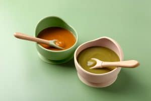 Green and pink baby bowls with pureed food and wooden-handled spoons on a light green surface; baby feeding products Australia.
